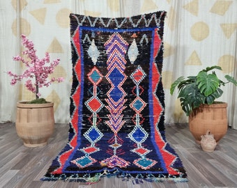 May include: A rectangular, hand-woven rug with a black background and geometric patterns in red, blue, and white. The rug features diamond and zigzag designs, with a fringe along the bottom edge. The rug is displayed on a wooden floor.