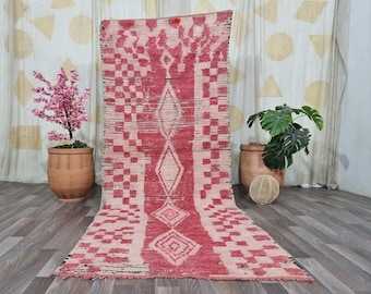 May include: A rectangular, hand-woven rug with a red and cream geometric pattern. The rug features diamond and square designs, with a faded, vintage aesthetic. The rug is displayed on a wooden floor, with decorative plants in the background.