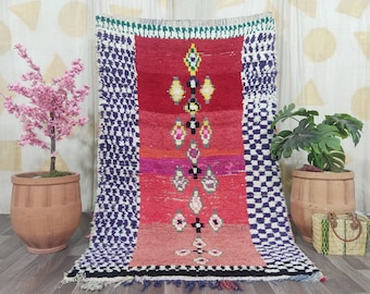 May include: A vibrant, rectangular rug with a central design in shades of red, pink, and orange, adorned with geometric patterns. The sides feature a checkered pattern in white and navy blue. The rug has a fringed edge.