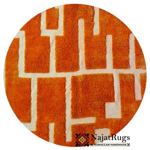 Puede incluir: Alfombra redonda naranja con un patrón geométrico blanco. La alfombra tiene una superficie texturizada y está etiquetada como "Najat Rugs Moroccan Handmade". La alfombra es circular y parece estar hecha de un material suave.