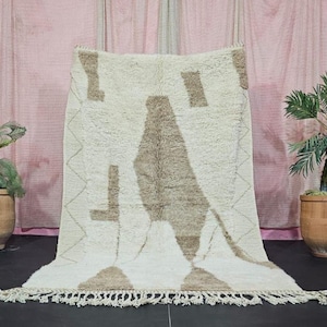 Könnte beinhalten: Ein cremefarbener Teppich mit geometrischem Muster und weicher, strukturierter Optik. Der Teppich hat ein großes, abstraktes Design in Braun- und Beigetönen mit Fransen am unteren Rand. Er wird vor einem rosa Hintergrund präsentiert.