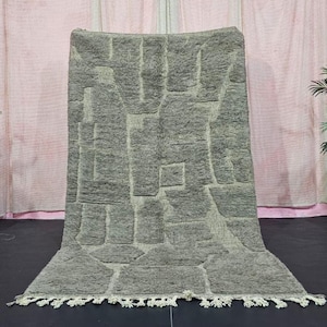 Könnte beinhalten: Ein grauer Teppich mit einem geometrischen Muster aus rechteckigen und quadratischen Formen. Der Teppich hat eine strukturierte Oberfläche und eine Fransen am unteren Rand. Der Teppich wird vor einem rosa Vorhanghintergrund präsentiert.