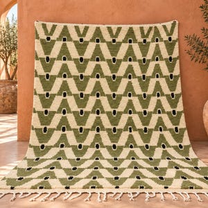 Puede incluir: Una alfombra rectangular con un patrón geométrico repetitivo de triángulos verde oliva y secciones blanco crema, acentuado con pequeñas formas de diamante negras. La alfombra está terminada con una fila de flecos blanco crema en el borde inferior.