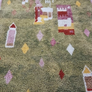 K&ouml;nnte beinhalten: Ein gr&uuml;ner Teppich mit einem farbenfrohen geometrischen Muster. Der Teppich hat einen Fransenrand und verf&uuml;gt &uuml;ber rosa, gelbe, rote und wei&szlig;e Akzente.