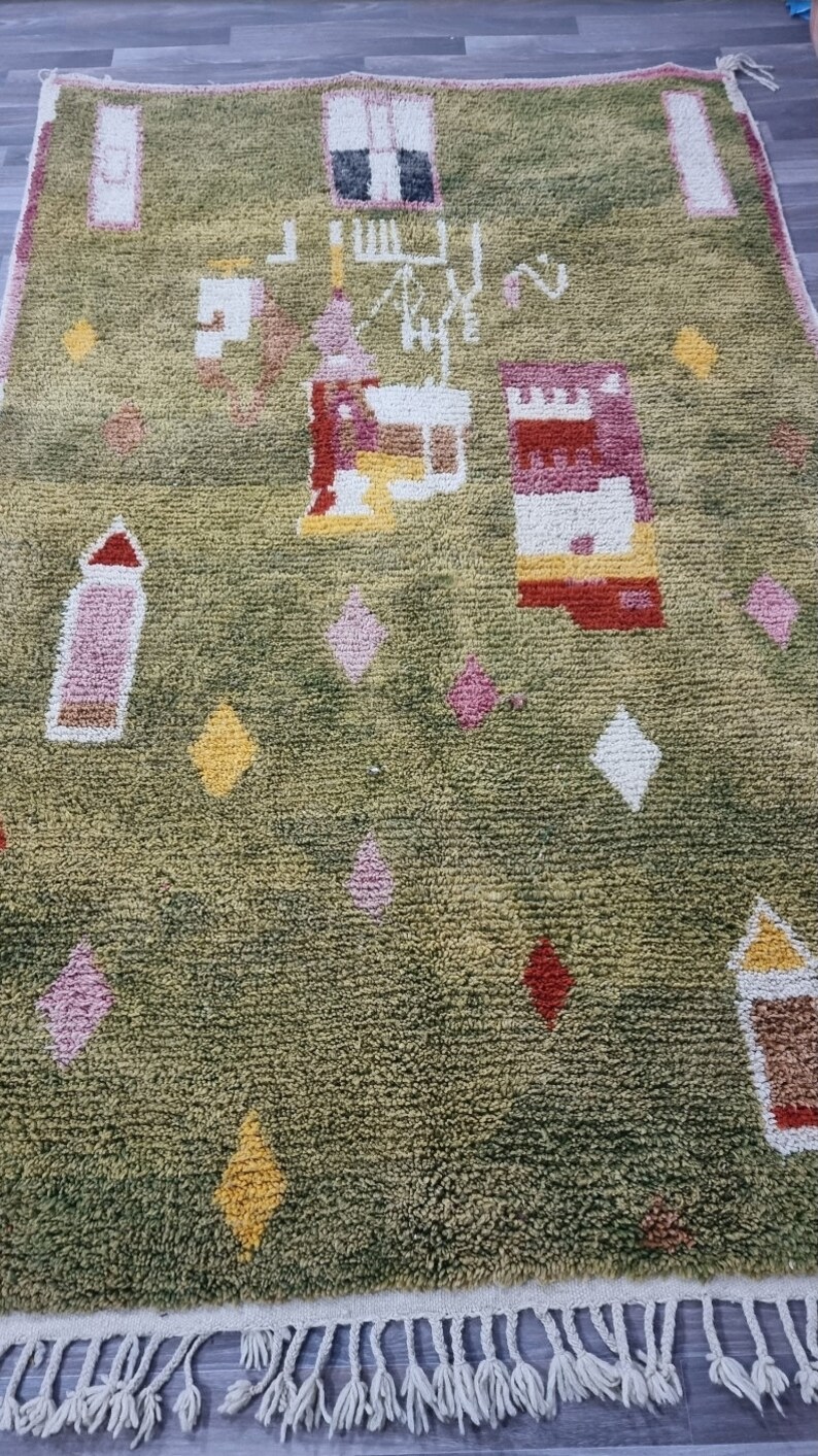 K&ouml;nnte beinhalten: Ein gr&uuml;ner Teppich mit einem farbenfrohen geometrischen Muster. Der Teppich hat einen Fransenrand und verf&uuml;gt &uuml;ber rosa, gelbe, rote und wei&szlig;e Akzente.