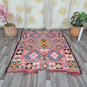 Pu&ograve; includere: Un tappeto rettangolare, tessuto a mano, con sfondo rosa e motivi geometrici arancioni, blu, bianchi e neri. Il tappeto ha un bordo nero e bordi sfrangiati. Il tappeto &egrave; posto su un pavimento in legno.