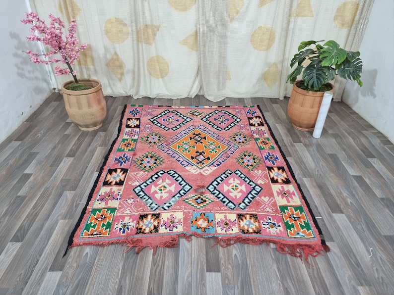 Pu&ograve; includere: Un tappeto rettangolare, tessuto a mano, con sfondo rosa e motivi geometrici arancioni, blu, bianchi e neri. Il tappeto ha un bordo nero e bordi sfrangiati. Il tappeto &egrave; posto su un pavimento in legno.