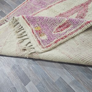 May include: A white and pink handwoven rug with a fringe border. The rug has a geometric pattern with pink and white stripes.