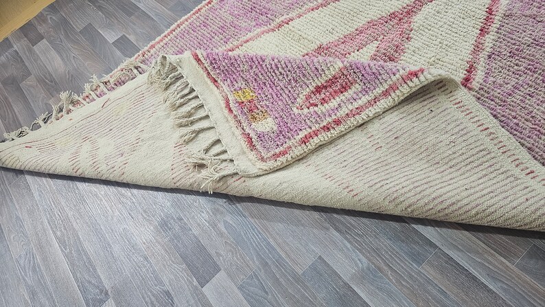 May include: A white and pink handwoven rug with a fringe border. The rug has a geometric pattern with pink and white stripes.