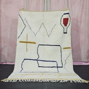 Könnte beinhalten: Ein cremefarbener, rechteckiger Teppich mit geometrischem Design. Der Teppich hat schwarze, blaue, goldene und graue Linien und Formen, darunter ein Kreuz, Zickzacklinien und eine flaschenartige Figur mit rotem Innenraum. Der Teppich hat einen Fransenrand.