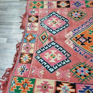 Pu&ograve; includere: Una sezione di un tappeto colorato e fantasia. Il tappeto presenta uno sfondo rosa con disegni geometrici in vari colori, tra cui blu, arancione, verde e nero. I bordi hanno una frangia.
