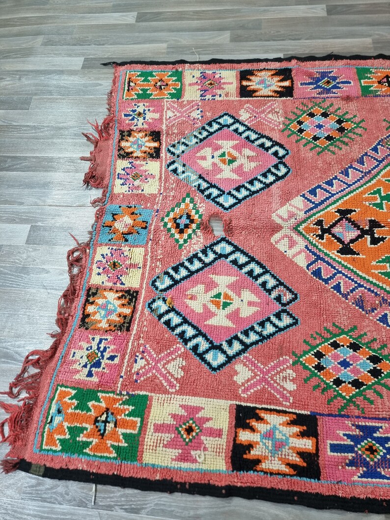 Pu&ograve; includere: Una sezione di un tappeto colorato e fantasia. Il tappeto presenta uno sfondo rosa con disegni geometrici in vari colori, tra cui blu, arancione, verde e nero. I bordi hanno una frangia.