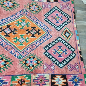 Pu&ograve; includere: Un tappeto rettangolare rosa con motivi geometrici arancioni, verdi, blu, neri e rosa. Il tappeto presenta forme a diamante e quadrate con disegni intricati e un bordo nero. Il tappeto &egrave; realizzato in materiale tessuto.