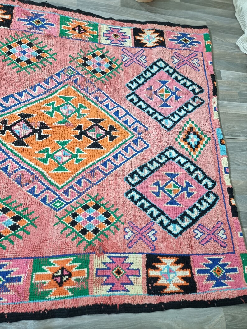 Pu&ograve; includere: Un tappeto rettangolare rosa con motivi geometrici arancioni, verdi, blu, neri e rosa. Il tappeto presenta forme a diamante e quadrate con disegni intricati e un bordo nero. Il tappeto &egrave; realizzato in materiale tessuto.