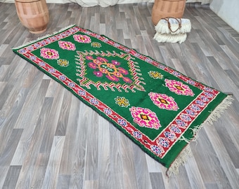 May include: A rectangular green rug with a central geometric design in pink, yellow, and orange. The rug features a red border with floral patterns and pink diamond-shaped motifs. Fringes are visible on the ends.
