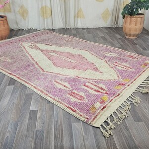 May include: A pink and white Moroccan rug with a diamond pattern and fringe. The rug is on a wooden floor.