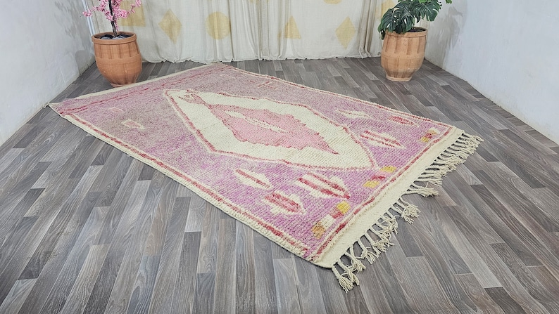 May include: A pink and white Moroccan rug with a diamond pattern and fringe. The rug is on a wooden floor.