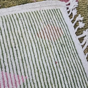 K&ouml;nnte beinhalten: Nahaufnahme eines wei&szlig;en und gr&uuml;nen gestreiften Teppichs mit Fransenrand. Der Teppich hat ein geometrisches Muster in Rosa und Braun.