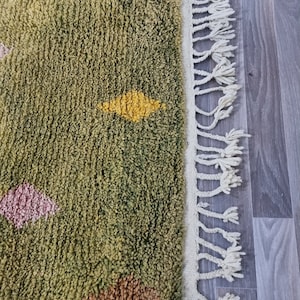 K&ouml;nnte beinhalten: Ein gr&uuml;ner Teppich mit einem wei&szlig;en Fransenrand. Der Teppich hat ein geometrisches Muster in verschiedenen Farben, darunter Gelb, Orange, Rosa und Braun.