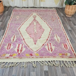 May include: A pink and white Moroccan rug with a diamond pattern and fringe. The rug has a traditional design with a central diamond shape and smaller diamond shapes around the edges.