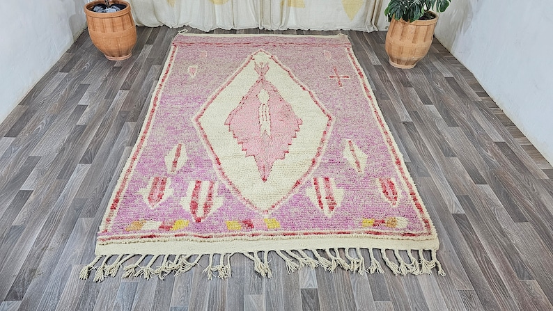 May include: A pink and white Moroccan rug with a diamond pattern and fringe. The rug has a traditional design with a central diamond shape and smaller diamond shapes around the edges.