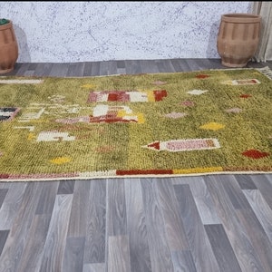 K&ouml;nnte beinhalten: Ein gr&uuml;ner, gelber, roter und wei&szlig;er marokkanischer Teppich mit geometrischem Muster. Der Teppich hat einen Fransenrand und liegt auf einem Holzboden.