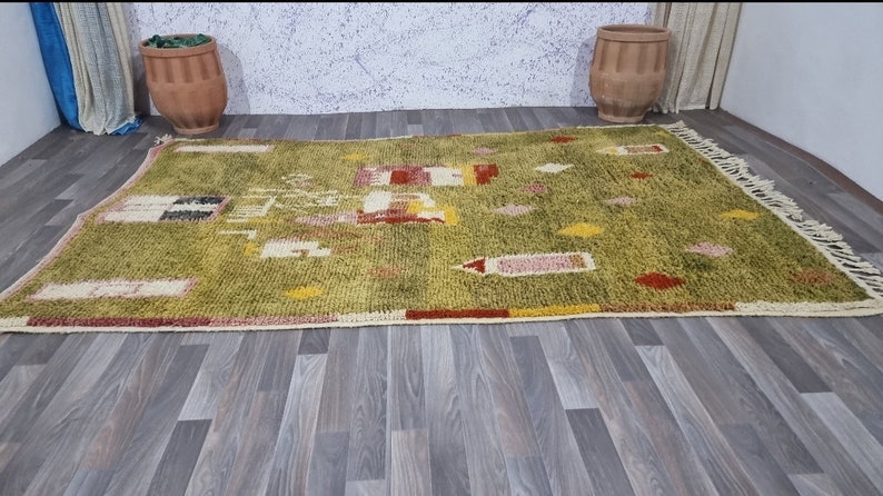 K&ouml;nnte beinhalten: Ein gr&uuml;ner, gelber, roter und wei&szlig;er marokkanischer Teppich mit geometrischem Muster. Der Teppich hat einen Fransenrand und liegt auf einem Holzboden.