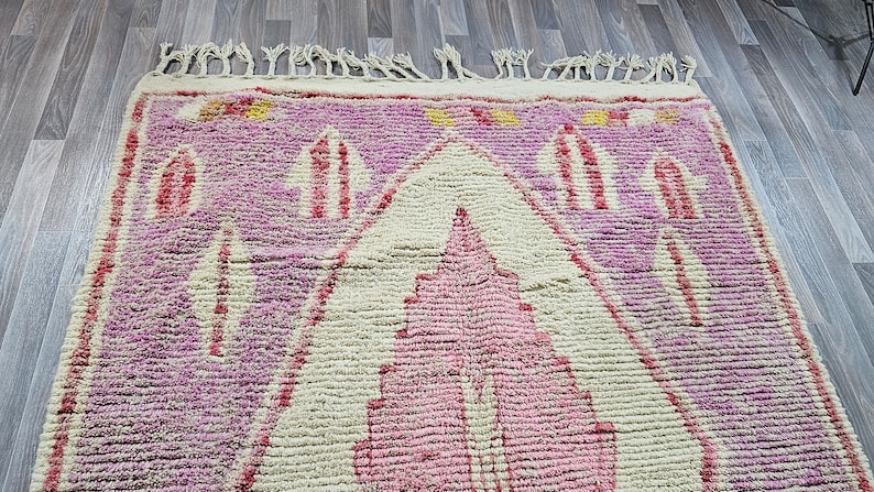 May include: A pink and cream Moroccan rug with a geometric pattern. The rug has a fringe border and is made of wool.