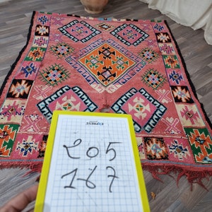 Pu&ograve; includere: Un tappeto rettangolare tessuto a mano con sfondo rosa e motivi geometrici arancioni, verdi, blu e neri. Il tappeto presenta motivi a rombi e quadrati. Le dimensioni sono 205 x 167 cm, come indicato su un blocco note giallo.