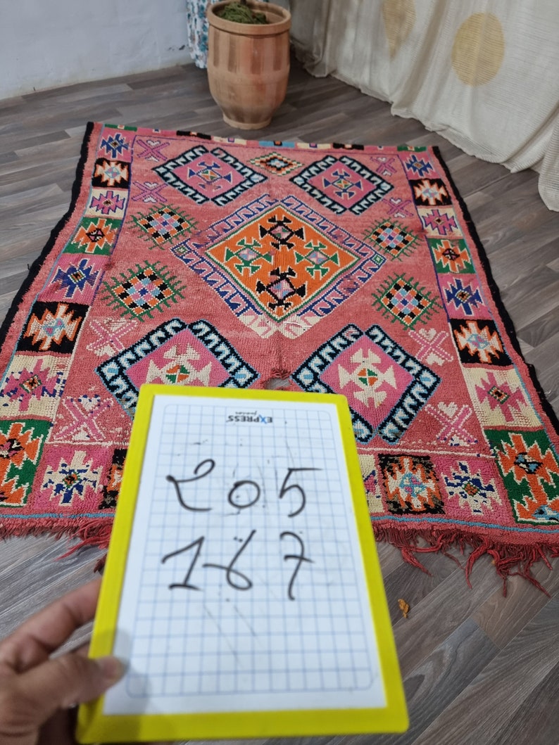 Pu&ograve; includere: Un tappeto rettangolare tessuto a mano con sfondo rosa e motivi geometrici arancioni, verdi, blu e neri. Il tappeto presenta motivi a rombi e quadrati. Le dimensioni sono 205 x 167 cm, come indicato su un blocco note giallo.
