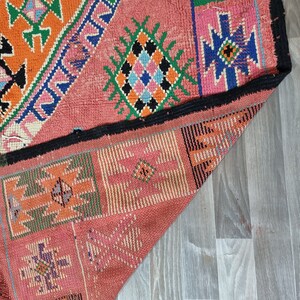 Pu&ograve; includere: Un tappeto colorato, tessuto a mano, con un design geometrico. Il tappeto presenta uno sfondo rosa con motivi geometrici arancioni, verdi, blu e neri. I bordi sono rifiniti in nero. Il tappeto &egrave; parzialmente piegato.