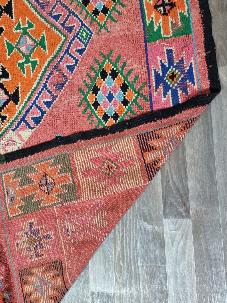 Pu&ograve; includere: Un tappeto colorato, tessuto a mano, con un design geometrico. Il tappeto presenta uno sfondo rosa con motivi geometrici arancioni, verdi, blu e neri. I bordi sono rifiniti in nero. Il tappeto &egrave; parzialmente piegato.