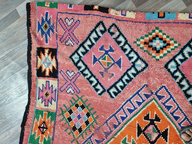 Pu&ograve; includere: Primo piano di un tappeto rosa con motivi geometrici blu, arancioni, verdi e neri. Il tappeto ha un bordo nero e una superficie strutturata.