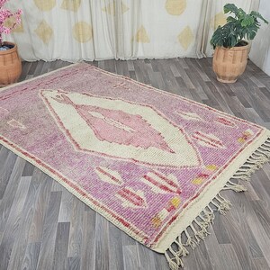 May include: A pink and white Moroccan rug with a diamond pattern and fringe. The rug is on a wooden floor.