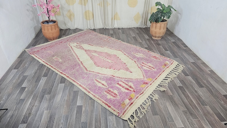 May include: A pink and white Moroccan rug with a diamond pattern and fringe. The rug is on a wooden floor.