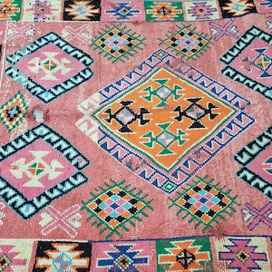 Pu&ograve; includere: Un tappeto rettangolare rosa con un motivo geometrico. Il tappeto presenta forme a diamante e quadrate in tonalit&agrave; di rosa, arancione, verde, blu e nero. Il tappeto ha un bordo nero ed &egrave; realizzato in materiale tessuto.