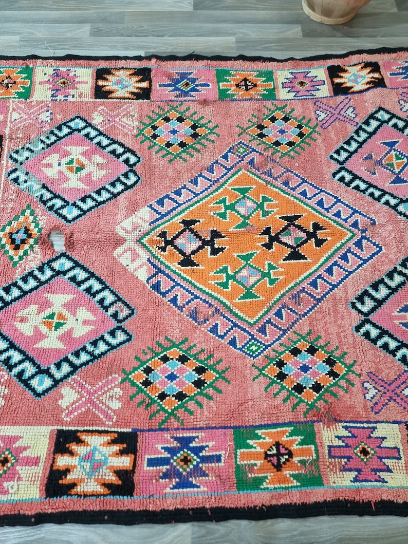 Pu&ograve; includere: Un tappeto rettangolare rosa con un motivo geometrico. Il tappeto presenta forme a diamante e quadrate in tonalit&agrave; di rosa, arancione, verde, blu e nero. Il tappeto ha un bordo nero ed &egrave; realizzato in materiale tessuto.