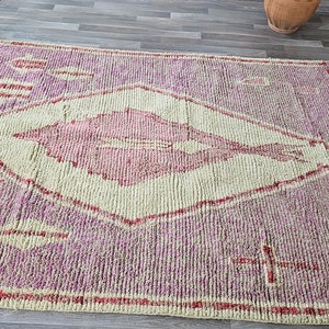 May include: A pink and white Moroccan rug with a diamond pattern and fringe. The rug has a traditional design with a central diamond shape and a repeating pattern of leaves.