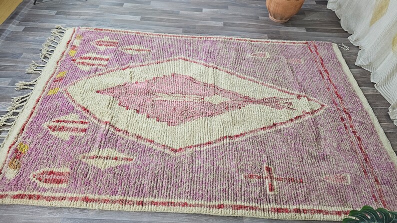 May include: A pink and white Moroccan rug with a diamond pattern and fringe. The rug has a traditional design with a central diamond shape and a repeating pattern of leaves.