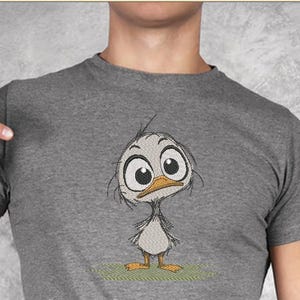 May include: A gray t-shirt featuring an embroidered cartoon duckling with large eyes and an orange beak. The duckling is standing on a patch of green grass. The shirt is a short-sleeved crew neck.