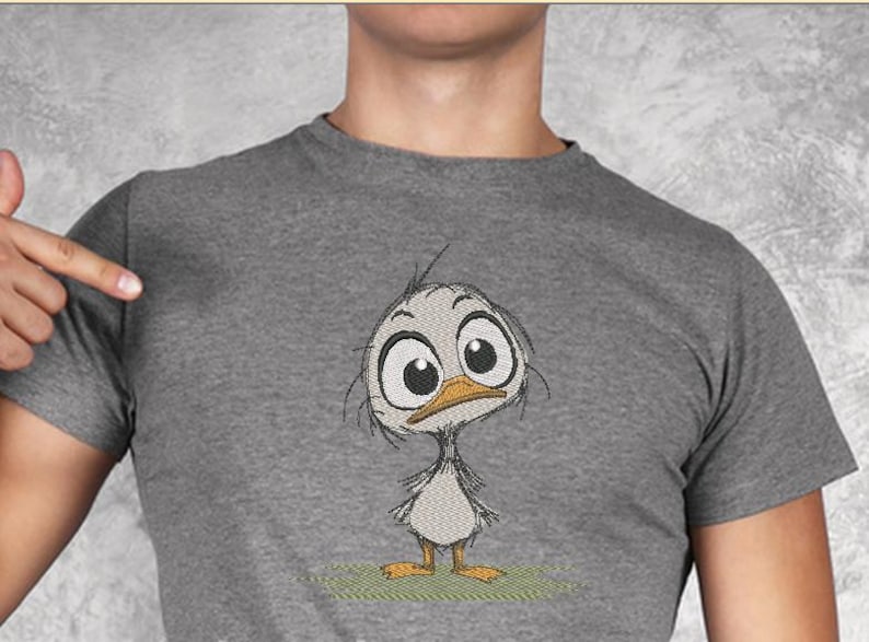 May include: A gray t-shirt featuring an embroidered cartoon duckling with large eyes and an orange beak. The duckling is standing on a patch of green grass. The shirt is a short-sleeved crew neck.