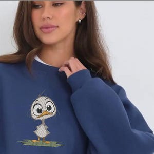 May include: Navy blue sweatshirt featuring an embroidered cartoon duck with large eyes and a surprised expression. The duck is standing on a patch of green grass. The sweatshirt has a crew neck and long sleeves.