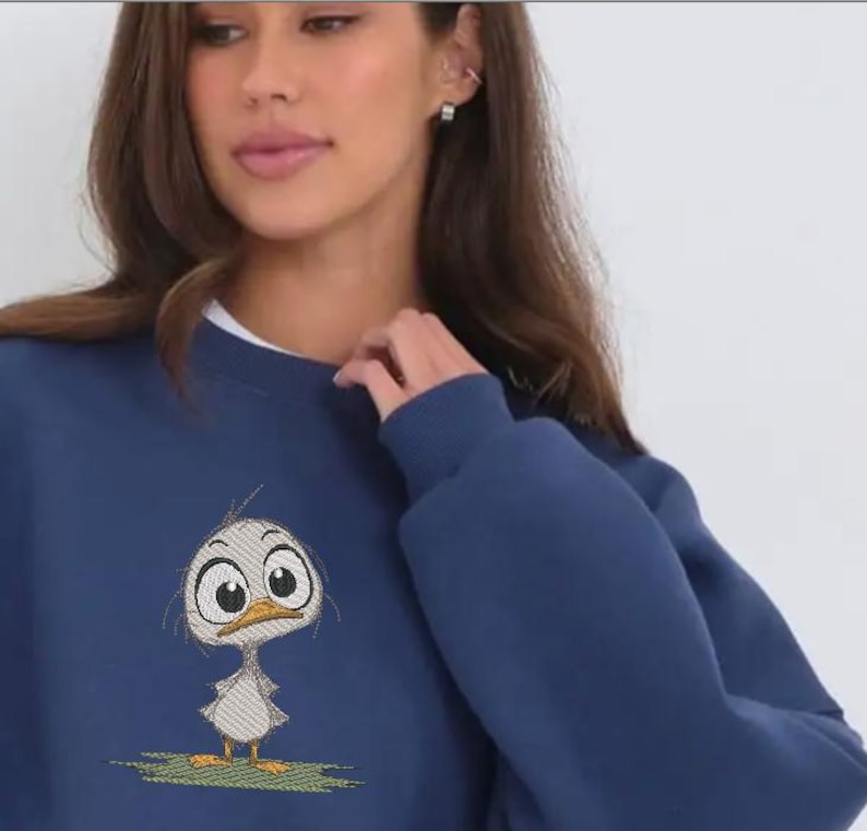 May include: Navy blue sweatshirt featuring an embroidered cartoon duck with large eyes and a surprised expression. The duck is standing on a patch of green grass. The sweatshirt has a crew neck and long sleeves.