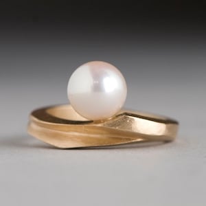 May include: A gold ring featuring a large, lustrous, white pearl. The ring has a unique, geometric design with a curved band. The pearl is centered on the ring, creating a classic and elegant look.