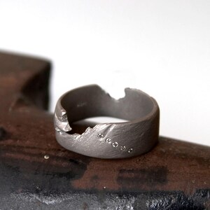 May include: A dark grey ring with a textured surface and an irregular edge. The ring has five small, clear stones set in a line. The ring is shown on a dark, textured surface.