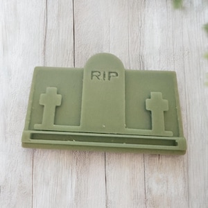 Puede incluir: Un objeto verde rectangular con forma de l&aacute;pida con las letras "RIP" talladas en la parte superior. Dos cruces est&aacute;n a cada lado de la l&aacute;pida. El objeto es probablemente un art&iacute;culo decorativo o un molde para manualidades.