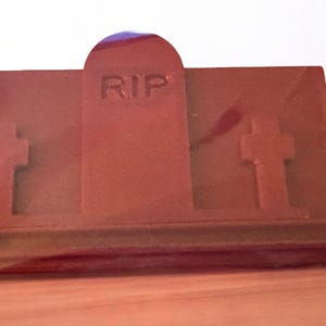 Puede incluir: Una l&aacute;pida marr&oacute;n con las letras "RIP" talladas en la parte delantera. La l&aacute;pida tiene dos cruces a cada lado y est&aacute; colocada sobre una superficie de madera.