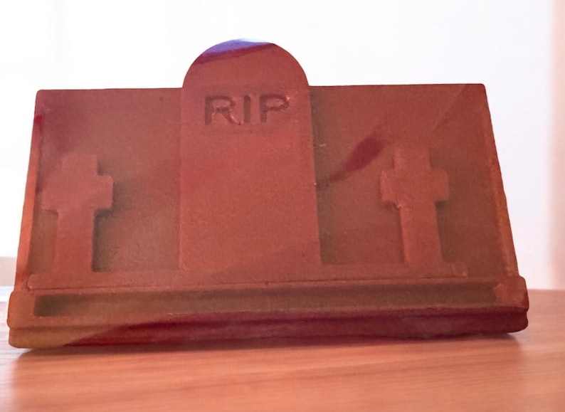 Puede incluir: Una l&aacute;pida marr&oacute;n con las letras "RIP" talladas en la parte delantera. La l&aacute;pida tiene dos cruces a cada lado y est&aacute; colocada sobre una superficie de madera.