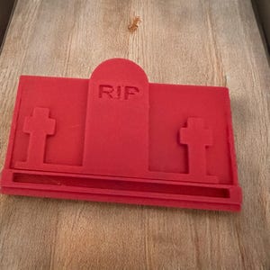 Puede incluir: Un objeto rojo brillante en forma de l&aacute;pida con las letras "RIP" talladas en la parte superior. Dos cruces est&aacute;n a cada lado de la l&aacute;pida. El objeto est&aacute; hecho de un material s&oacute;lido.