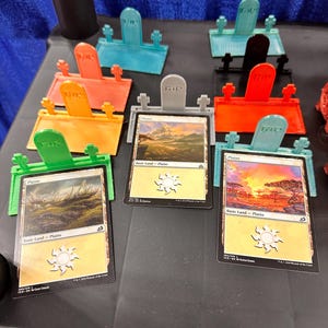 May include: A collection of colorful, 3D-printed card holders shaped like tombstones, each holding a trading card. The cards are labeled "Plains" and feature landscape artwork. The tombstones are in various colors, including orange, green, and blue.